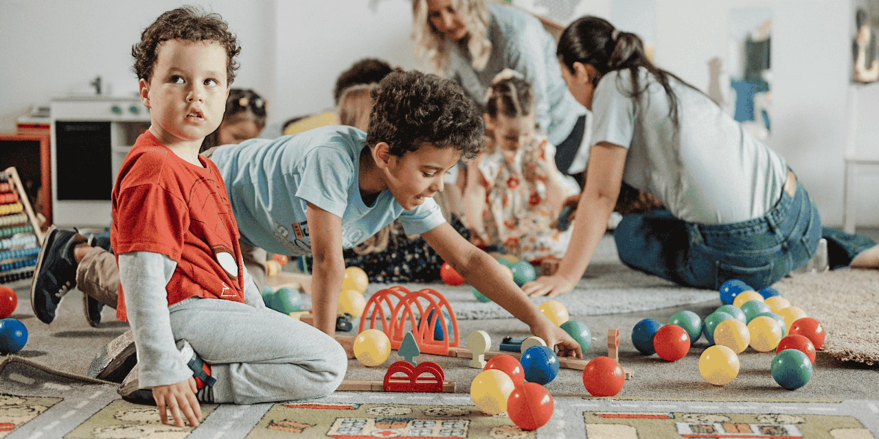 Children playing with colorful toys on a carpet, assisted by adults in the background.